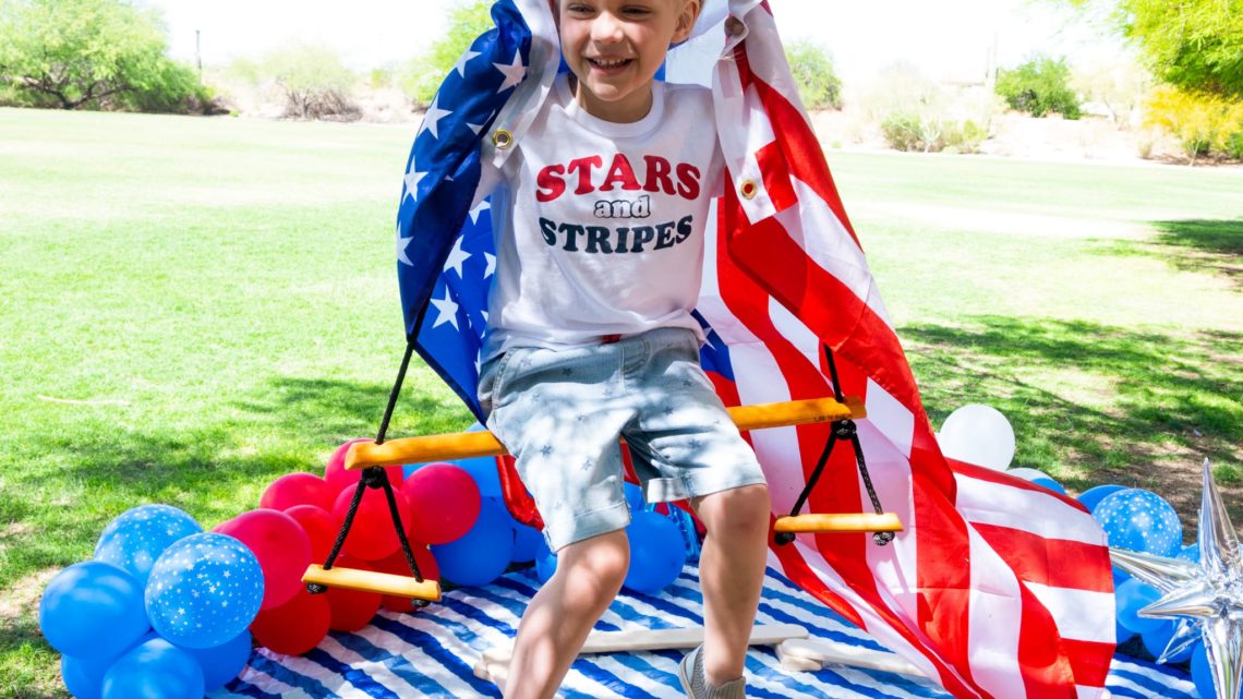 Happy child on a swing holding an American flag behind her, with red, white, and blue balloons on the ground