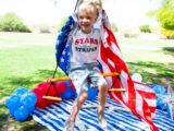 Happy child on a swing holding an American flag behind her, with red, white, and blue balloons on the ground